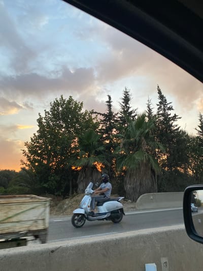 A person rides a white scooter on a highway at dusk. The sky is filled with dramatic clouds, transitioning from a bright orange on the left to pale blues and grays on the right. A line of trees, including palm trees and evergreens, forms the background. In the foreground, a blurred trailer is visible on the left, and the side mirror of a car, also showing a blurred car on the highway, is on the right. The scooter rider is wearing a helmet and a gray shirt, and is moving from left to right. The highway has a concrete barrier. The scene appears to be in El Karia, Algeria.
