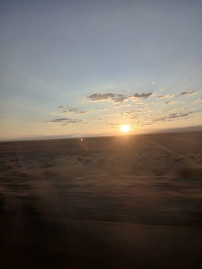 The image captures a sunset over a flat, arid landscape, possibly the steppes surrounding Uzynbulak, Kazakhstan. The sun, a bright, diffused orb, is positioned low on the horizon, casting a warm glow across the sky and illuminating scattered clouds. The sky transitions from a pale blue at the top to a soft yellow and orange near the horizon. The foreground is a blur of brown and tan, suggesting movement, likely from a vehicle. In the midground, low-lying vegetation or dry grasses cover the terrain, appearing slightly blurred due to motion. A faint horizontal line, possibly a power line or a distant ridge, is visible across the landscape. The overall impression is one of a vast, open space at dusk.
