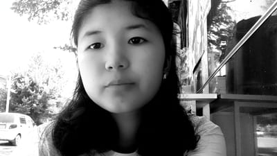 A black and white, close-up selfie shows a young East Asian woman outdoors. She has dark hair, bangs, and is wearing a white t-shirt and a small stud earring in her visible ear. The background includes foliage and trees, a white car, and what appears to be a storefront or building with reflective surfaces. The lighting suggests it is daytime. The image is a selfie taken from the chest up, focusing on the woman's face.