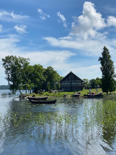 The image depicts a tranquil scene at a lakeside village, likely in Patanevshchina, Russia, as suggested by the architectural style and natural landscape. A traditional wooden house with a steep roof sits on a grassy embankment, surrounded by lush green trees. Several wooden rowboats are moored along the shore, some partially submerged in the clear water. Reeds and aquatic plants grow in abundance along the water's edge, their reflections shimmering on the surface. The sky above is a vibrant blue, dotted with fluffy white clouds, suggesting a pleasant, sunny day. A few individuals are visible near the house and boats, engaged in seemingly casual activities, adding a touch of human presence to the idyllic setting. The overall impression is one of a peaceful, rural existence, deeply connected to nature.