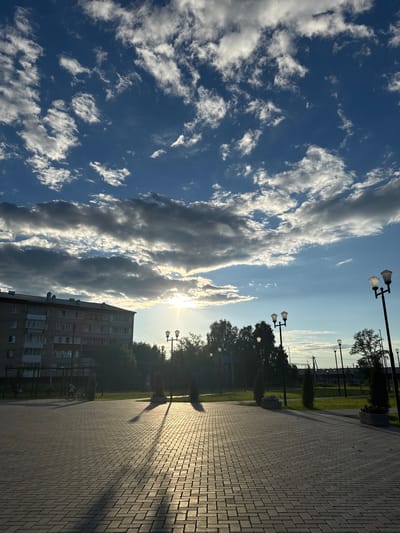 The image captures a sun-drenched plaza with a patterned brick walkway, bordered by grass and trees. In the background, a multi-story apartment building stands to the left, its facade illuminated by the setting sun. Several lampposts, typical of urban street lighting, are strategically placed throughout the scene, casting long shadows on the walkway. The sky above is a mix of blue and white clouds, with the sun peeking through, creating a dramatic interplay of light and shadow. The overall atmosphere is serene, suggesting a quiet evening in the city of Mozhga, Russia.