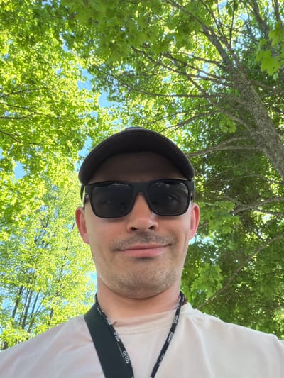 A man wearing a black flat cap and sunglasses is facing the camera. He also wears a white shirt and a black lanyard with "FOX40" printed on it. The camera angle is looking upwards at the man, with green trees and a clear blue sky visible in the background. The location is outdoors, with the trees and sky suggesting it could be a park or wooded area. The image is likely taken in Oreshniki, Russia, due to context. The man appears to be taking a selfie.