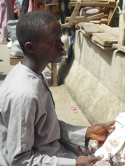 In Fagge D1, Nigeria, a young male with short dark hair and dark complexion is depicted in right profile, seated outdoors. He wears a light grey long-sleeved tunic. His hands, positioned in the lower right frame, hold a white rectangular retail package containing multiple small, clear, spherical items with a yellowish-orange tint, consistent with decorative beads or earrings. The ground is light-colored and appears dusty. To the subject's right and behind him, a low, light grey concrete or plastered wall runs horizontally, topped by several rough wooden planks. Further into the background, elements of an outdoor market are visible, including wooden support structures, textiles with patterns, and the partial view of another individual wearing light pink attire. A white bag rests on a wooden pallet behind the subject to the left. The scene is illuminated by natural daylight.