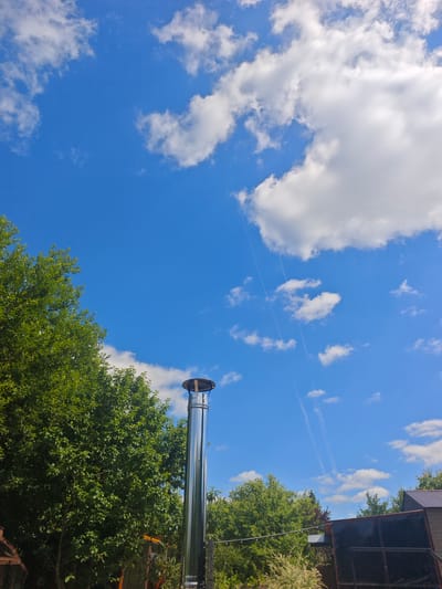 This media file depicts a bright, sunny day in a rural outdoor setting, likely in Khlevino, Russia, given the provided context. The primary subjects are a tall, reflective metal chimney and lush green trees. The chimney stands prominently in the foreground, reaching towards a vibrant blue sky filled with scattered white clouds. The clouds exhibit some wispy, elongated trails, suggesting the recent passage of an aircraft.
The scene is bathed in clear daylight, indicating it is likely mid-morning or early afternoon. The weather appears to be fair and warm. The background shows more foliage from trees and a glimpse of a dark-colored roofline of a building, hinting at a residential or agricultural property. The overall impression is one of a tranquil, possibly summer day in the countryside. There is no visible text in the image. The composition focuses on the interplay between the man-made structure (chimney) and the natural elements (sky, trees), with the bright sunlight casting a cheerful mood.