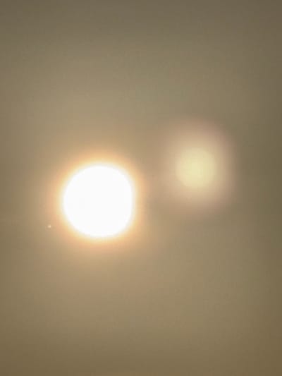 The image depicts a hazy atmospheric scene, stated to be in Kato Karioutes, Greece. Two luminous, circular objects are visible against a uniform, light brown-beige background. The primary light source is located in the lower-left quadrant, appearing as a bright white core surrounded by a distinct yellow-orange halo. To its upper-right, a secondary, less intense and more diffuse light source is present, characterized by a soft, yellowish-white glow with indistinct edges. The background exhibits a consistent, grainy texture, suggesting a high level of atmospheric diffusion or haze. No individuals, animals, structures, vegetation, or discernible ground features are present. No actions or interactions are depicted.