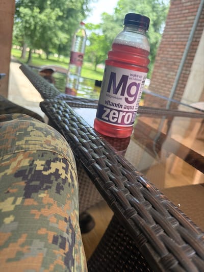 A plastic bottle of Magnesium Vitamin Aqua Zero drink rests on a brown wicker table. Camouflage patterned clothing is visible in the lower right of the image. The background shows trees and a brick building. There is no indication that this image was taken in Dolna Mitropolia, Bulgaria.