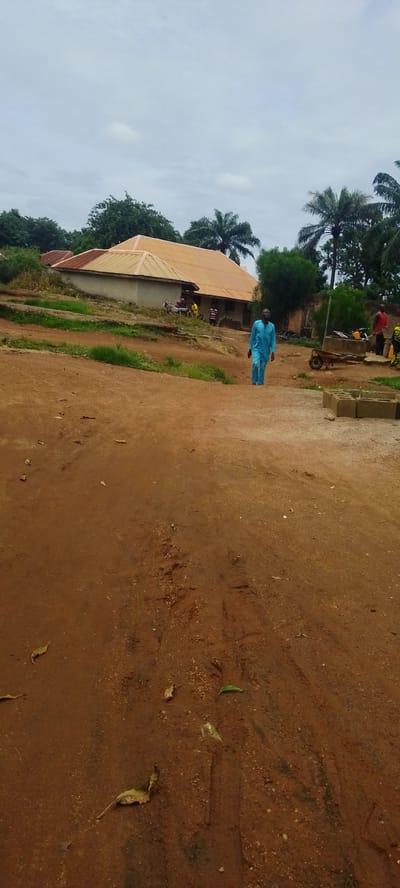 The image depicts an outdoor scene in Baissa, Nigeria, likely during the daytime under an overcast sky, suggesting it might be cloudy or nearing dusk. The ground is predominantly reddish-brown dirt, with some patches of dry grass and fallen leaves scattered throughout. In the background, a building with a distinctive orange or copper-colored roof is visible. It appears to be a single-story structure with lighter-colored walls.

Several people are present in the scene. One man, wearing a bright blue traditional outfit, is walking towards the viewer on the dirt path. Further back, near the building, a few other individuals are gathered, though their features are less distinct. One person in a yellow shirt and another in a striped shirt are visible. To the right of the building, there's a wheelbarrow and what looks like a motorcycle or motorbike parked. A few stacks of what appear to be concrete blocks are also visible on the right side of the frame.

The overall setting suggests a rural or semi-rural environment in Nigeria. The architecture of the building, the clothing of the people, and the presence of elements like the wheelbarrow and the motorbike are consistent with this location context. The sky is a pale gray, indicating diffused light, and there are no strong shadows, reinforcing the idea of an overcast or cloudy day. There is no visible text in the image.