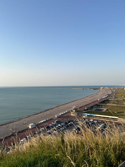 This is a high-angle, landscape-oriented photograph taken from a vantage point overlooking a coastal scene in Dieppe, France. The image is rotated 90 degrees clockwise.
The scene depicts a wide, pebbly beach bordering a calm, blue-grey sea. The shoreline stretches along the bottom of the frame, with a paved promenade running parallel to it. Numerous people are visible scattered along the beach and promenade, enjoying the outdoors. A long pier or breakwater extends out into the sea from the middle of the beach.
Adjacent to the promenade is a long, busy parking lot filled with a variety of cars. Beyond the parking lot, there is a strip of green grass and some modern-looking buildings, possibly part of a resort or leisure facility. In the far distance, the coastline continues, with a hint of a town or city visible on the horizon.
The sky is a clear, soft blue, transitioning to a lighter shade near the horizon, suggesting it is late afternoon or early evening. The overall atmosphere appears peaceful and leisurely, indicative of a pleasant day by the sea. Tall, dry grasses in the foreground frame the right side of the image, adding a natural element and a sense of depth. No text is discernible in the image.