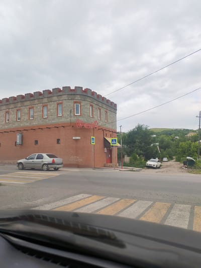 The image shows a street scene in Volsk, Russia, featuring a building with a castle-like facade, a pedestrian crossing, and several vehicles. The building, constructed with red brick and stone, has crenellated battlements along the top and rows of windows. A neon sign, partially obscured, is mounted on the corner of the building. A silver sedan is driving across a pedestrian crossing in the foreground, while a white SUV and another white car are parked further down a dirt road in the background. Green trees and a gently rolling hill are visible beyond the buildings. The sky is overcast with scattered birds. The overall impression is a somewhat rural or suburban setting within the city.