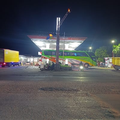 The image depicts a nighttime scene at a "PERTAMINA" gas station in Kecamatan Asembagus, Indonesia, viewed from a road with a rough, asphalt surface. The gas station canopy is brightly illuminated, standing out against the dark sky.
In the center of the frame, a large, green and orange double-decker bus, possibly belonging to "Bintang Jaya" (text partially visible), is parked at the gas pumps. Several individuals are gathered near the bus on the left, some sitting beside a small, three-wheeled vehicle. On the far left, a yellow delivery truck is in motion, appearing blurred due to its movement. Another yellow truck is partially visible on the right. A person on a motorcycle with its headlight on is positioned in the mid-ground, near the bus.
The activity suggests a stop for refueling or a break from travel. People are seen resting or waiting, indicating a transient moment during a journey. The scene is lit by various artificial sources, including the gas station lights, streetlights, and vehicle headlights, creating pockets of brightness amidst the darkness. A tall light pole with two rectangular lights and an orange signal light is prominent in the foreground. Trees with visible foliage are discernible on the right side, illuminated by ambient light.
