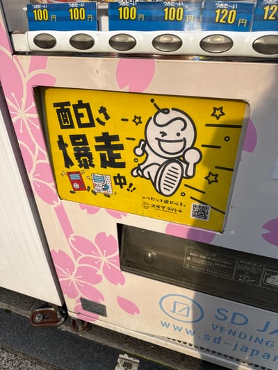 The image shows a close-up of a vending machine in Toshima, Japan, with a pink cherry blossom motif on its side. Prominently displayed on the front is a yellow sticker with black Japanese text and a cartoon drawing of a happy character running. The text on the sticker reads "面白く走って〜〜〜!" (Omoshiroku hashitte! - Run amusingly!) and below it, "いっだって遊びをも。" (Ittatte asobi o mo. - Let's play anytime.). There are also smaller illustrations of two red and blue robots on the sticker, and a QR code. Above the yellow sticker, price tags are visible, showing prices in Japanese Yen (円). One tag reads "100円" (100 Yen) and another reads "120円" (120 Yen). The lower part of the image reveals a black slot for dispensing items and some warning instructions in Japanese. The ground in front of the vending machine appears to be asphalt with some debris.