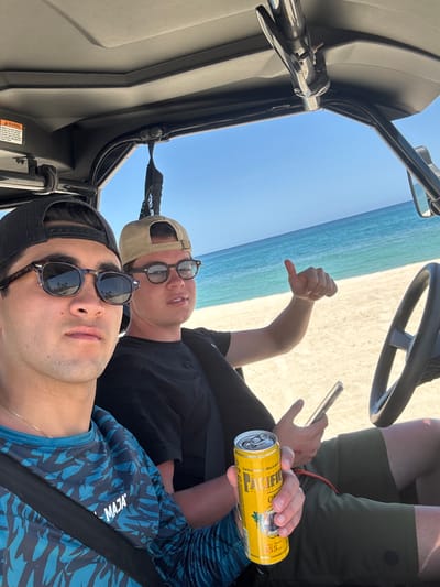 Two men are in an off-road vehicle. The location appears to be a beach in La Capilla, Mexico. The man in the foreground is wearing a blue shirt with a camouflage pattern, a black cap, and sunglasses. He is also wearing a seatbelt. The man in the background is wearing a black shirt, khaki cap, and glasses, and gives a thumbs-down. He is holding a cell phone in his left hand and a can of "Pacifico" beer in his right hand. The vehicle's interior is visible, as is the sky and sandy beach.