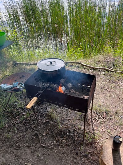 This media file depicts an outdoor scene with a barbecue grill and food being cooked. The setting appears to be a natural environment, with tall green reeds or grass growing alongside what could be a body of water. The ground is covered with dirt, dry leaves, and twigs, suggesting a wooded or semi-wild area.
The main object of focus is a metal barbecue grill with a fire burning underneath. Several items wrapped in foil, likely food, are placed on a rack above the flames, and smoke is rising from the grill. A large metal pot with a lid sits on the grill, obscuring some of the food. A metal grate is positioned on top of the grill to hold the pot. A wooden handle with the word "CAMPING" visible is attached to the grill. There is also a rusty circular metal surface on the ground near the grill, possibly another cooking surface or a fire pit lid. In the background, a black can with a silver lid is perched on a log. A bright green bucket is visible in the lower left corner.
The image is taken from a slightly elevated, angled perspective, with the scene oriented vertically. The lighting suggests it is daytime, with sunlight casting shadows. The overall mood is casual and perhaps related to an outdoor activity like camping or picnicking. The location context provided, Вяззеўскі сельскі Савет, Belarus, further reinforces the idea of a rural or natural setting. There are no people visible in the image.