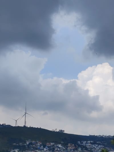 The image shows a cloudy sky over Đa Thọ, Vietnam. Two wind turbines stand on a hill overlooking a residential area of small buildings. The sky is mostly covered in gray and white clouds, with patches of blue visible.