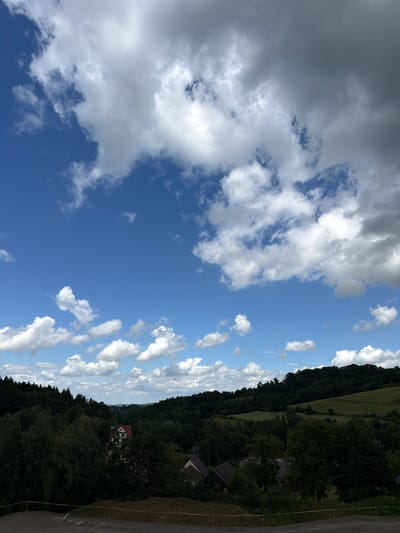 The image depicts a picturesque landscape under a partly cloudy sky. In the foreground, there is a cleared area with some construction materials and yellow caution tape, suggesting an ongoing development project. Beyond this, a dense forest of dark green trees dominates the mid-ground, interspersed with a few houses with red roofs, characteristic of rural architecture. In the distance, rolling hills covered in lush vegetation extend towards the horizon. The sky is a vibrant blue, adorned with a mix of fluffy white cumulus clouds and darker, more ominous storm clouds, hinting at changing weather conditions. The overall scene conveys a sense of natural beauty and rural tranquility, with a subtle indication of human activity. This scene is located in the vicinity of Jodłownik, Poland.