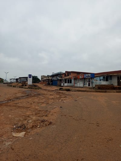 The scene depicts a street in Oshogbo, Nigeria on an overcast day. The road is unpaved and heavily eroded with large potholes. Along the street are a series of small shops and stalls, some open and some closed. A few cars are parked or moving in the distance. A sign stands near the edge of the road. The overall impression is one of a poor area with infrastructure challenges.
