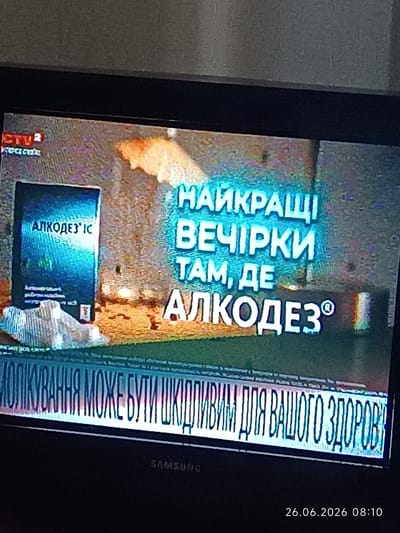 The image shows a television screen displaying what appears to be a news or advertisement segment. The text on the screen is in Ukrainian. The top left corner has a logo that reads "ETV" followed by some Ukrainian text. In the center of the screen, there is a large blue book-like object with the text "АЛКОДЕЗ'ІС" on it. To the right of this, there is glowing text that reads "НАЙКРАЩІ ВЕЧІРКИ ТАМ, ДЕ АЛКОДЕЗ". Below this, a scrolling banner displays the text "МОЛІКУВАННЯ МОЖЕ БУТИ ШКІДЛИВИМ ДЛЯ ВАШОГО ЗДОРОВ'Я". At the bottom right of the screen, a timestamp is visible: "26.06.2026 08:10". The background of the screen appears to be a textured, abstract design with orange and yellow hues. A "SAMSUNG" logo is visible on the bezel of the TV at the bottom. There is no identifiable location within the city of Bolhrad, Ukraine, depicted in this image; it is a captured image of a television screen.