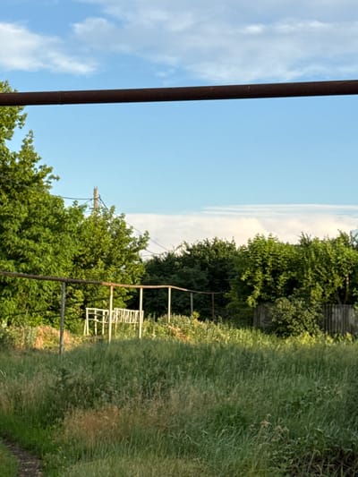 The image depicts an outdoor scene in a grassy area, possibly a yard or field, with a clear blue sky and scattered clouds. A thick, rusty metal pipe runs horizontally across the upper portion of the frame. In the middle ground, a structure made of white metal, possibly a swing set or a frame, is visible amidst overgrown green vegetation. Trees with lush green foliage are abundant, lining the background and to the left of the frame. A weathered wooden fence is also present on the right side. A dirt path can be seen in the lower left corner. The image appears to be taken in Rossosh, Russia, during daylight hours, suggesting a temperate climate. There are no people or animals visible in the scene. The overall impression is one of a rural or semi-rural setting with elements of both nature and man-made structures.