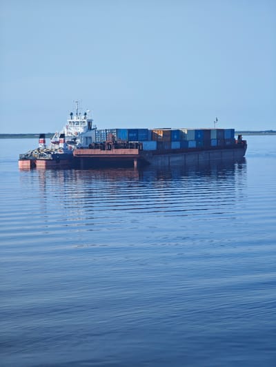 A large barge is being pushed by a tugboat on a calm body of water. The barge is loaded with numerous shipping containers, stacked several high. The tugboat is white with red and black accents and has various antennas and equipment on its deck. The barge itself is a rusty brown color. In the distance, a low-lying landmass with vegetation is visible under a clear, pale blue sky. The water is a deep blue and reflects the sky and the vessels, with gentle ripples on its surface. The scene appears to be set during the day, with bright, even lighting suggesting it could be midday or early afternoon. The water and sky suggest a peaceful and perhaps cold environment, consistent with a northern location like Dudinka, Russia, which is known for its Arctic climate and port. No people are clearly visible on the vessels. There is some text on the shipping containers, including "SNCOR" and "OCL," but it is largely unreadable due to distance and resolution. The overall impression is one of industrial transport in a natural, serene setting.