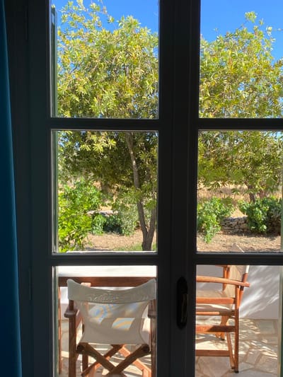The image displays an outdoor scene captured from within a shadowed room through an open, light-framed French door system. The setting is Πλατύς Γιαλός, Greece.
The foreground is dominated by the light gray (or white) wooden frame of the French doors, which are divided into six panes by vertical and horizontal mullions. A dark, vertical door handle is visible on the central vertical mullion. To the far left, a portion of a blue curtain is visible.
Through the lower panes, an outdoor patio is visible. The patio floor is paved with irregular, light-colored flagstones. Two wooden-framed outdoor chairs are present: one with a white canvas backrest is partially visible on the left, oriented slightly towards the viewer, with distinct patches of sunlight exhibiting minor chromatic aberration on its fabric. A wooden table is partially obscured behind it. Another similar chair is positioned on the right. A white wall forms the right boundary of the patio.
Through the upper and middle panes, a natural landscape is visible under a clear, bright blue sky. The scene is dominated by multiple trees with abundant, light-green foliage. The ground beneath the trees appears dry and earthy, interspersed with patches of lighter, drier vegetation. Dry-stacked stone walls are discernible in the mid-ground among the trees and greenery. The overall environment is brightly illuminated by direct sunlight, casting distinct shadows. No individuals are present in the scene.