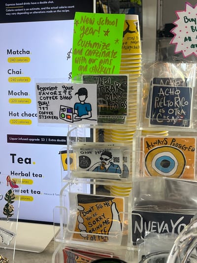 The image depicts an interior view of a 787 Coffee shop in El Paso, United States, centered on a merchandise display and a digital menu board. On the left, a white digital menu displays beverage options such as Matcha (240 calories), Chai (240 calories), Mocha (320 calories), Hot Chocolate (320 calories), and Tea, with an option for a $3 liquor infused upgrade.

Adjacent to the menu, a clear acrylic multi-tiered display rack holds various stickers and pins, individually packaged in clear plastic sleeves. At the top of this display, a fluorescent green handwritten sign states, "New School year! Customize and caffeinate with our pins and stickers!". Behind this sign, a stack of yellow disposable coffee cups with black graphics is visible.

Merchandise on the display includes:
*   A white sticker depicting a cartoon person in a baseball cap using a laptop next to a 787 Coffee logo with colored squares, bearing the text "Represent Your FAVORITE COFFEE SHOP Buy! 787 COFFEE STICKERS."
*   A dark sticker with white graffiti-style text "DEBI TIRAR MAS FOTOS."
*   A blue sticker with white text "ACHO PUERTO RICO ES OTRA COSA" and a small Puerto Rican flag.
*   A white sticker with "ONE HAND" and "787 COFFEE CO" logos, featuring a cartoon man in sunglasses drinking from a coffee cup.
*   An orange sticker with an "evil eye" symbol and the text "ALWAYS PROTECTED."
*   An orange sticker depicting a hand holding a coffee cup with the text "DON'T TELL ME YOU'RE SORRY BRING ME COFFEE."
*   A partially visible dark sticker at the bottom right reads "NUEVA YO[RK]."

On the far left, below the digital menu, a separate display stand holds small metal pins, including a red high-heeled shoe and two gold-colored leaf designs. A pink starburst-shaped sign with white polka dots and partially visible text beginning "Buy B[UY] ALL..." is located on the far right of the scene. No individuals are depicted.