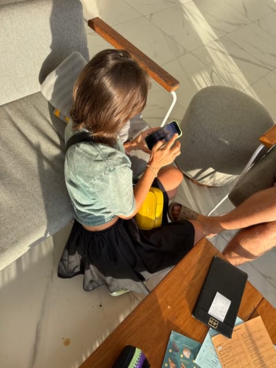 Captured from an elevated perspective, the scene unfolds within a restaurant setting, featuring a woman seated on a gray cushioned chair, engrossed with her smartphone. Her attire consists of a green top and a black skirt, complemented by a yellow fanny pack. Adjacent to her sits a man, his bare legs visible, adorned with tan sandals.

A wooden table stands between them, hosting a black bill folder, menus, and other indiscernible objects. Sunlight streams into the location, casting shadows across the marble floor. The scene likely occurs within Kedisan, Indonesia, as indicated by the OCR text extract from the menu.