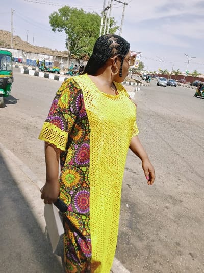 The image displays an outdoor scene on a paved road in Communauté rurale de Payar, Senegal, under clear daylight. A woman, positioned in the foreground, is partially turned away from the camera, looking towards the right. She has braided black hair styled with intricate patterns on the side, wears large gold hoop earrings, and light-framed eyeglasses. Her attire is a long dress, vertically divided, with one side composed of a bright yellow open-knit fabric and the other side featuring a dark fabric patterned with vibrant, multicolored concentric circles. She holds a dark object in her left hand; her right hand exhibits pink nail polish. The background reveals a multi-lane asphalt road with a black and white striped curb or median. Various vehicles, including a green auto-rickshaw on the immediate left, other smaller vehicles, and multiple cars, are present on the road. A low concrete wall separates the road from a rocky hillside and scattered green trees. Utility poles and streetlights are also visible against the sky.