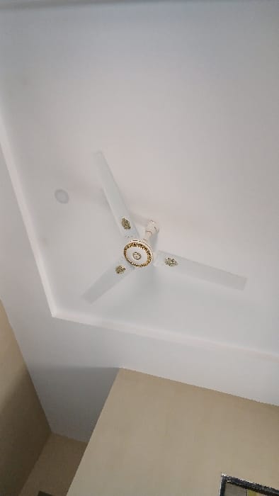 An upward-angled view captures the interior of a room in Bashar, Nigeria. A three-bladed ceiling fan, white with gold-colored decorative accents, is centrally mounted on a white, false ceiling with a recessed border detail. To the upper left of the fan, a single white recessed light fixture is visible in the ceiling. The walls in the lower portions of the frame are painted a light beige. A partial view of a dark-framed opening, possibly a window or door, is visible in the bottom right corner of the image. No people are visible. The fan is stationary.