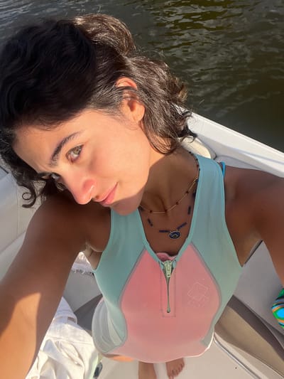 A young adult female is depicted in a close-up, high-angle shot aboard a white boat. She has dark, wavy hair and light-colored eyes. Her complexion appears tanned, with direct sunlight illuminating her face and casting shadows. She wears a sleeveless, two-tone garment, likely a wetsuit or rashguard, featuring a light blue upper section and a light pink main body with a central zipper and a subtle geometric logo. Two layered necklaces are visible: a gold chain with small red beads and a longer necklace with blue beads and an oval blue pendant. The subject is looking towards the camera. Behind her, calm, dark water is visible, indicating the boat is on a river or lake. This scene occurs on the Minho River, adjacent to the city of Vila Nova de Cerveira, Portugal.