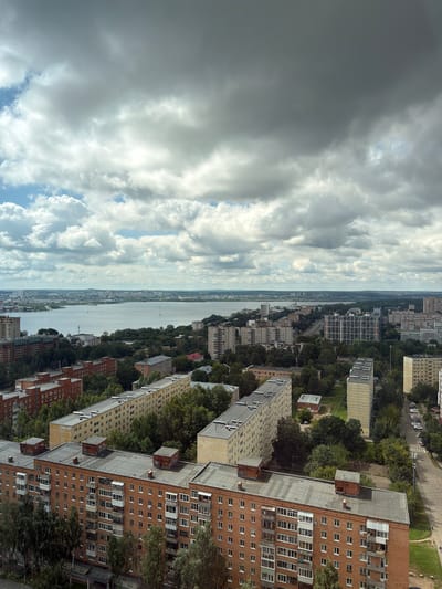 The image captures a wide-angle view of a city with a large body of water in the background. The sky is predominantly filled with heavy, dark clouds, with patches of lighter clouds and some blue sky peeking through.
In the foreground and midground, there are numerous apartment buildings. The buildings are of various heights and architectural styles, common in Soviet-era urban planning. Many are multi-story, with consistent window patterns and balcony structures. Some buildings are brick-faced, while others appear to be concrete or stucco. Lush green trees are interspersed among the buildings, suggesting a well-established urban green space.
The water in the background appears to be a wide river or a large lake. Along its banks, there are more buildings, some of which appear to be industrial or commercial, and a distant cityscape with a hazy horizon. There are some structures that might be bridges or infrastructure crossing the water or along its edges.
The lighting suggests it is daytime, but the cloud cover creates a diffused, somewhat somber atmosphere. There are no visible people or vehicles in the immediate foreground, but distant elements suggest urban activity. The overall impression is of a densely populated urban environment with a significant natural water feature. The location is identified as Verkhnii Liuk, Russia.