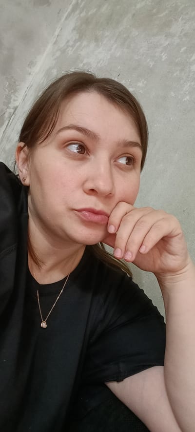 A young woman with light brown hair, wearing a black shirt and a delicate necklace, is pictured indoors against a textured gray wall. Her right hand rests on her chin, and she gazes to her right. The lighting suggests an indoor setting, possibly a home. The location is specified as Хохлы, Russia.