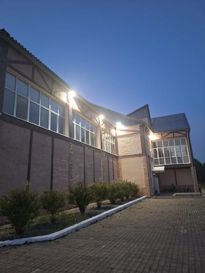 The image captures a building with a rustic architectural style, characterized by its textured, light-colored walls and dark wooden beams. The building features large, multi-paned windows that reflect the twilight sky. Outdoor lights illuminate the facade, casting a warm glow. In the foreground, a paved pathway made of interlocking bricks leads towards the building. A white-painted curb separates the pathway from a row of neatly trimmed bushes. Beyond the bushes, the ground is covered with grass and small yellow flowers. The sky above is a clear, deep blue, indicative of early evening. In the distance, at the entrance of the building, two individuals can be seen, one standing and another seated, suggesting the presence of activity or a gathering. The overall scene depicts an exterior view of a building in what appears to be a park or public area in Атамановка, Russia, during the twilight hours.