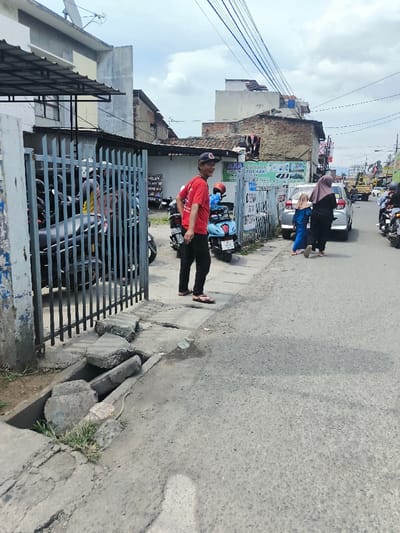 The image captures a bustling street scene on what appears to be a cloudy but bright day. The perspective is from the side of the road, looking down a street lined with buildings and vehicles.
In the immediate foreground on the left, a broken concrete drainage ditch or culvert runs parallel to the road, with some small patches of grass and weeds growing from it. The pavement next to it is uneven and worn.
A dark metal gate, possibly part of a business or residential entrance, stands partially open on the left. Through the gate, several motorcycles, primarily scooters in various dark colors, are parked. Behind these vehicles, parts of buildings are visible, including one with a flat roof and a satellite dish.
On the pavement to the right of the gate, a man is standing. He is wearing a red short-sleeved t-shirt, black pants, and brown flip-flops, along with a dark baseball cap. He has a slight beard and is looking towards the viewer with a friendly smile. Behind him, a light blue scooter is parked.
Further down the street on the right side, two individuals are walking away from the camera. One is a taller figure, likely a woman, wearing a dark hijab and dark clothing. Beside her, a smaller figure, possibly a child, is dressed in blue. A silver car is either parked or slowly moving behind them. More motorcycles are visible further down the street, and a yellow construction vehicle or truck can be seen in the distance on the right.
Along the street, various buildings rise, some appearing quite old with exposed brickwork or aged concrete. Overhead, a dense network of black power lines spans across the street against the backdrop of an overcast sky filled with white clouds.
Visible text includes:
* On a blue sign or banner attached to a wall behind the man in red: "AMA Collection" is clearly visible on the top line, followed by "GROSIR KAOS KAKI" which translates to "Wholesale Socks." Images of socks are also present on this sign.
* A partially obscured white sign further down the street shows the word "DITERIMA," meaning "Accepted" or "Received" in Indonesian.