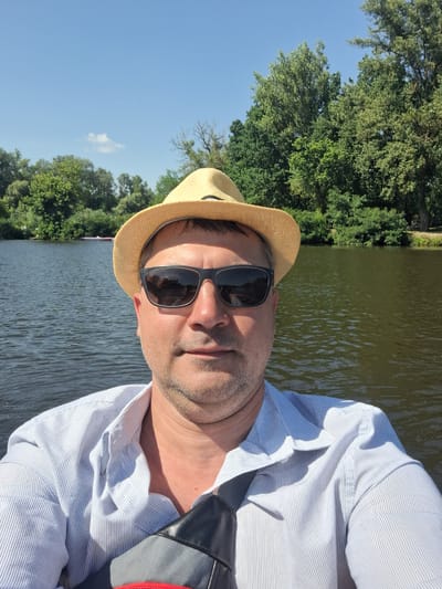 The image is a selfie of a man in his 40s, wearing a straw hat and sunglasses, taken from the chest up. He is on a body of water, likely a river, with trees and a clear blue sky in the background. The man is wearing a light blue and white striped shirt. He appears to be outdoors, possibly on a boat, given the water's surface and the presence of vegetation along the bank. The location is identified as Potoky, Ukraine. The water is rippling slightly, and sunlight reflects off its surface. The trees are lush and green, suggesting a warm weather setting.
