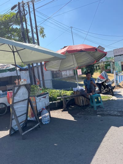 In Sumokali, Indonesia, on a bright, sunny day, a street vendor sits on a turquoise plastic stool, looking at his phone. He is seated on a wooden bench under a large red umbrella, next to a motorcycle covered with a tarp. In front of him, a stall is set up with a white display board advertising prices in Indonesian Rupiah and a stack of green limes. A large bottle of drinking water sits on the ground near the stall. The street is paved with asphalt, and a sidewalk is visible to the right. Overhead, a complex network of power lines crisscrosses the clear blue sky.