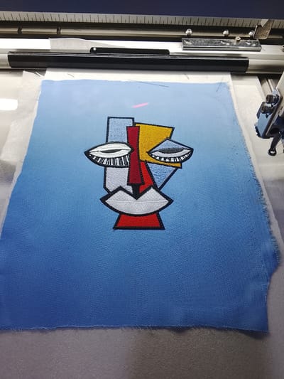 The image shows a piece of blue fabric with an embroidered abstract face design. The face is made up of geometric shapes and lines in various colors, including red, yellow, white, and black. The embroidery is detailed, with thin black outlines defining the shapes and intricate stitching for features like the eyes and lips. The fabric itself has a subtle gradient, transitioning from a lighter blue at the top to a darker blue at the bottom. The edges of the fabric are raw and appear to be unfinished, suggesting it's part of a larger project or a sample. In the background, parts of what appears to be an embroidery machine are visible, with metallic components and a guiding ruler. The overall impression is of a close-up view of a piece of textile art in progress, likely in a workshop or studio setting. There is no visible text in the image.