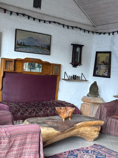 The image depicts an interior scene, likely a living room, filled with rustic and vintage decor. Dominating the foreground is a large, organic-shaped wooden coffee table with a rough-hewn texture. On this table sits a carved wooden bowl. To the left, partially visible, is an armchair draped with a pink and striped textile. Behind the armchair is a bed with a deep red headboard, covered by an intricately patterned rug.
The background reveals a white brick wall adorned with several decorative elements. A large, framed landscape painting hangs on the wall above the bed, depicting a mountain range, a river, and a village. To the right of this painting, a dark wooden grandfather clock with a visible clock face is mounted on the wall. Below the clock, a small wooden shelf holds several small metallic cups or votives. Further to the right, another framed painting, this one depicting a snowy landscape with trees, is displayed. Below this painting, a bronze sculpture of a hooded figure holding a child rests on a multi-tiered wooden pedestal.
Decorative string lights with colored bulbs are strung along the upper part of the white wall and across the angled ceiling, adding a festive or ambient touch. The flooring appears to be tiled. The overall impression is one of a cozy, eclectic space with a focus on natural materials and antique pieces. There is no discernible connection to Alagir, Russia, in the image itself; the description is purely based on the visual elements present.