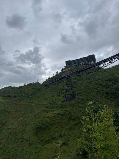 The image shows a hillside in Cat Cat Village, Vietnam, on a cloudy day. The hillside is covered in dense green vegetation. A large, abandoned metal structure, possibly an unfinished building or transport system, runs along the side of the hill. The sky is overcast with gray clouds. A few branches of foliage are visible in the upper right corner. The overall scene appears somewhat desolate.