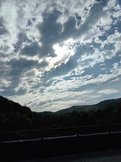 The scene depicts a landscape in Hòa Hiệp, Vietnam, under a dynamic sky. The upper two-thirds of the image are dominated by a mixture of grey stratocumulus and white cumulus clouds. The sun is obscured by a dense cloud mass in the upper-left quadrant, producing a distinct bright halo of light. Below the sky, a continuous line of dark green, densely forested hills or mountains extends horizontally across the midground. The foreground consists of a heavily shadowed, dark barrier structure, resembling a concrete guardrail, positioned above a dark, undifferentiated surface, likely a road or path. No individuals, actions, or interactions are visible.