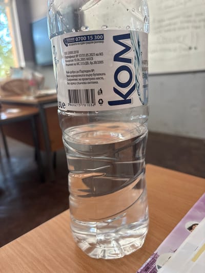 A clear plastic bottle of water, labeled "KOM," is placed on a light brown wooden table. The label features Bulgarian text, a phone number "0700 15 300," and standard product information like ISO 22000:2018 certification and a barcode. The bottle contains clear water, and droplets of condensation are visible on its surface. In the background, out of focus, a classroom or office environment is suggested by furniture and possibly a whiteboard or screen. The overall scene is static and indoors. The location is the city of Dimovo, Bulgaria.