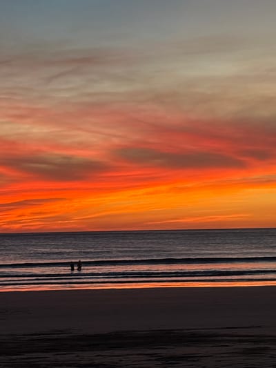 The image captures a sunset at a beach in Cabo Velas, Costa Rica. The sky is filled with a dramatic mix of colors ranging from light grays and whites to vibrant oranges and reds, indicating the setting sun. At the edge of the beach, a body of water reflects the sky's orange hues. Two figures are visible in the water, appearing as small silhouettes. The coastline transitions from a wet, reflective surface to a darker, sandy section.