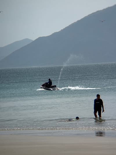 The image depicts a serene coastal scene with a person on a jet ski in the middle ground, creating a spray of water. In the foreground, two individuals are wading in the shallow water near the shore, with one person standing and the other appearing to be swimming or playing. The backdrop features rolling hills or mountains under a hazy sky, characteristic of the landscape near Dong Ninh Hoa, Vietnam. The water is calm with gentle ripples, and the beach in the immediate foreground is composed of light-colored sand. The overall atmosphere is one of leisure and outdoor recreation.