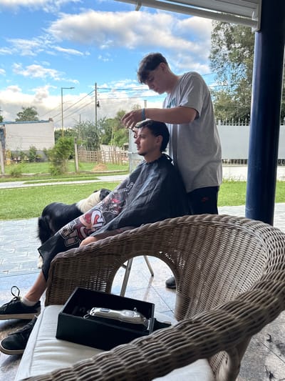 This image depicts an outdoor haircutting scene taking place on a covered patio or veranda in El Hueco, Argentina. Two males are present: one seated, receiving a haircut, and another standing behind him, performing the haircut.
The seated male, with dark hair and a mustache, wears a black barber cape adorned with graphic designs featuring text and skull imagery. He is dressed in dark shorts and black sneakers with white socks. He sits in a light-colored wicker chair with a white cushion, facing right, with his profile visible.
The standing male, positioned behind the seated individual, wears a grey short-sleeved t-shirt and dark shorts or pants displaying a red Nike swoosh logo. He has a light blue wristband on his right wrist. He holds a pair of scissors in his right hand and a comb in his left, actively trimming the hair of the seated man.
On the white cushion of a partially visible wicker chair in the foreground, a silver hair clipper is resting inside an open black rectangular case. Small dark fragments, consistent with cut hair, are visible on the tiled ground near this chair.
A black and white dog, possibly a Border Collie, is visible on the green grass immediately behind the seated man.
The environment behind the patio features a well-maintained green lawn, followed by a wooden fence, various trees and bushes, and distant residential structures with white walls and tiled roofs. Utility poles with overhead wires and a street light are also discernible in the background. The sky above is blue with scattered white clouds, indicating daylight conditions. The patio itself has a white overhead cover and a dark blue supporting pillar on the right side of the frame. The ground beneath the patio is tiled.