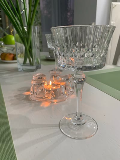 This image captures a still life composition on a white table, possibly in a dining or living area. The primary subjects are various glass objects. A large, intricately patterned crystal wine glass is positioned horizontally, its bowl facing left. Next to it, also on its side, are two small, faceted glass tea light holders, each containing a lit candle. The warm glow from the candles illuminates the surrounding glass surfaces and casts soft shadows on the table. In the background, a clear glass vase filled with long, green stems of flowers is visible. The stems suggest a bouquet, possibly decorative. A hint of fruit, including what appears to be a green apple and a reddish-brown fruit (perhaps an apple or pear), can be seen within the vase, adding a touch of color to the arrangement. A textured green placemat or runner is partially visible along the bottom and right edge of the frame. The lighting suggests it is evening or indoors, with artificial light from the candles being the dominant source. The overall mood is tranquil and elegant, emphasizing the interplay of light and glass. There is no visible text in the image.