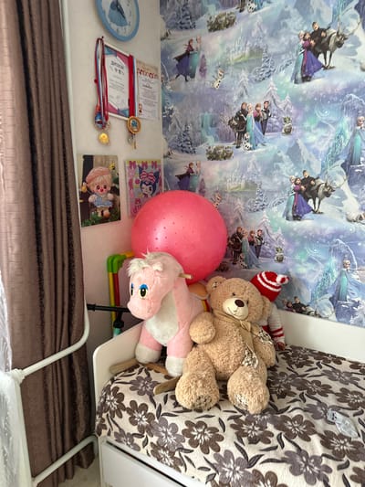 The image depicts a child's bedroom, likely in a residential setting in Мариинский, Russia. The room is decorated with "Frozen" themed wallpaper, featuring characters from the movie in a snowy landscape. On the wall, there is a light blue clock and a framed diploma with medals attached. Several plush toys are present: a pink rocking horse, a large teddy bear wearing a red and white striped hat and scarf, and a smaller plush toy is partially visible on the bed. A large, pink exercise ball is also visible. The bed is covered with a floral patterned blanket. A brown curtain hangs on the left side of the image, and a white metal bed frame is visible. The overall impression is that of a well-loved child's room, filled with toys and personal items.
