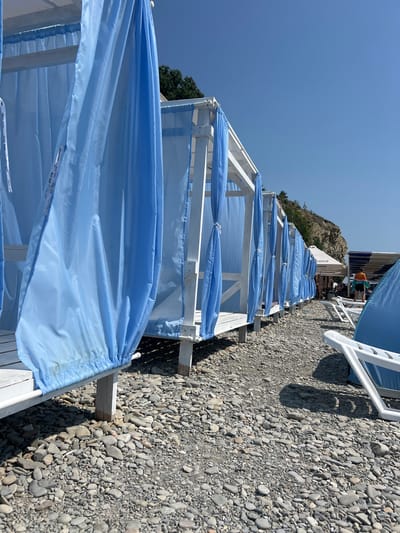 On a sunny day, a row of white wooden beach cabanas with blue curtains is visible on a pebbled beach. The cabanas are arranged in a line, receding into the distance. The pebbled ground is covered with smooth, gray stones of various sizes. In the background, a steep, rocky cliff with sparse vegetation rises against a clear blue sky. Beyond the cliff, a small structure with a white tent and some striped awnings is visible, along with a few people. A white plastic lounge chair is positioned in the foreground on the right, partially obscuring a blue tent. The overall scene depicts a resort area on the coast of Blagoveshchenskaya, Russia.