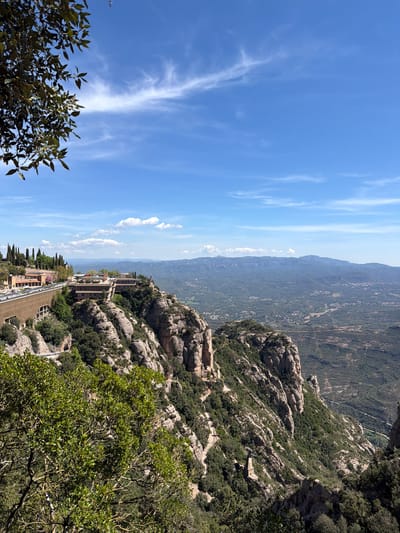 This image captures a panoramic view of the Montserrat mountain range in Collbató, Spain, under a bright, clear blue sky with sparse, wispy cirrus clouds. The scene is observed from a high vantage point, looking out over a dramatic landscape characterized by distinctive, jagged rock formations covered with dense green Mediterranean vegetation.
In the midground, several modern buildings with multi-level terraces and flat roofs are integrated into the steep, rocky slopes, suggesting a monastery complex or visitor facilities. Winding paths or roads are visible along the mountainside, connecting these structures. The foreground features more of the characteristic rocky terrain and lush green trees, with some foliage partially framing the bottom left and right of the view. In the far distance, a hazy horizon line separates the sky from further undulating mountain ranges and valleys.
The bright sunlight suggests midday or early afternoon, casting distinct shadows and highlighting the rugged textures of the rocks. The weather is clear and sunny. There are no people visible in the image, nor is any discernible text present. The overall impression is one of grandeur, natural beauty, and the serene integration of human structures within a powerful natural environment.