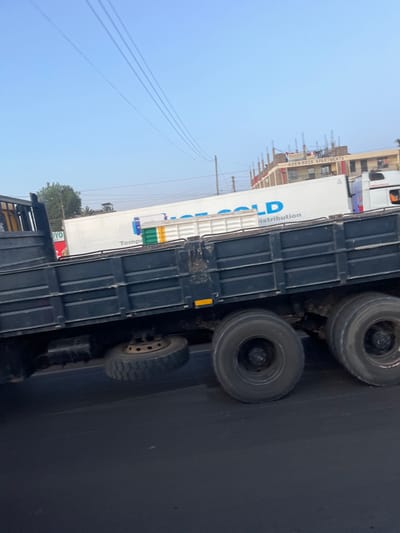 The image displays an urban street scene in Biashara ward, Kenya, under a clear blue sky. In the foreground, a dark-colored flatbed truck is positioned on an asphalt road, with its rear wheels visible. A spare tire is mounted beneath the truck's chassis, visible between the axles. Behind this truck, a white semi-trailer truck with "HOT COLD Distribution" branding on its side is partially visible, followed by the cab of another white semi-trailer truck displaying "ADEN TRUCK" on its side. In the background on the right, a multi-story building, identified by a sign as "ADEN ROCK APARTMENTS," shows visible construction elements on its upper levels. Electrical power lines traverse the upper portion of the frame. No individuals are discernible in the scene.