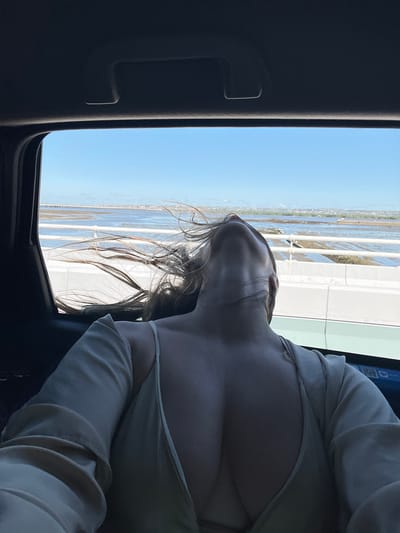 The image shows a person's back and head tilted back, with their hair blowing in the wind, seen through the window of a vehicle. The person is wearing a light-colored dress with a low neckline. Outside the window, there is a body of water with a long pier or bridge in the distance. The sky is clear and blue. The scene appears to be a coastal area. The city of South Denpasar is located in Indonesia, but the image does not provide any specific visual cues to confirm this location.