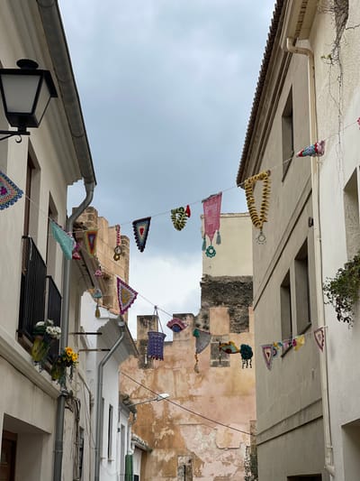 The image displays a narrow street or alleyway in Buñol, Spain, flanked by multi-story buildings under an overcast sky. The street is festively decorated with numerous multi-colored crocheted items strung between the buildings at various heights. These decorations consist of diverse shapes including triangular flags, squares, circular motifs, bell-shaped elements, and a distinctive long, coiled yellow piece, all exhibiting intricate patterns and vibrant colors such as pink, teal, purple, yellow, and green.
On the left side, a light-colored building features a black, ornate street lamp mounted high on the wall, a metallic rain gutter and downspout system, and a window with a black metal balcony railing. Two green hanging containers beneath the balcony hold artificial white and yellow flowers.
The building on the right, also light-colored with a slightly darker tiled roof edge, has a vertical downspout and a vine growing down its wall. Windows with dark frames are visible on both structures.
In the background, at the terminus of the street, a weathered, light reddish-brown stone wall, indicative of an ancient fortification or castle, rises prominently. This structure presents sections of darker stone and visible crenellations. The sky is uniformly grey and cloudy, providing diffuse light to the scene. No individuals are present in the depicted area.